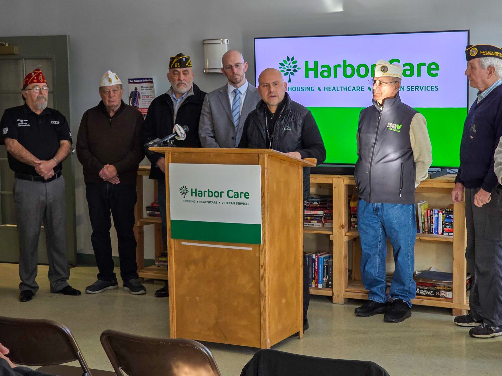 A group of men stand around a podium with a Harbor Care sign in the background. Some wear veteran hats with insignias. One man speaks into a microphone, while others listen attentively. Shelves with books are visible in the background.