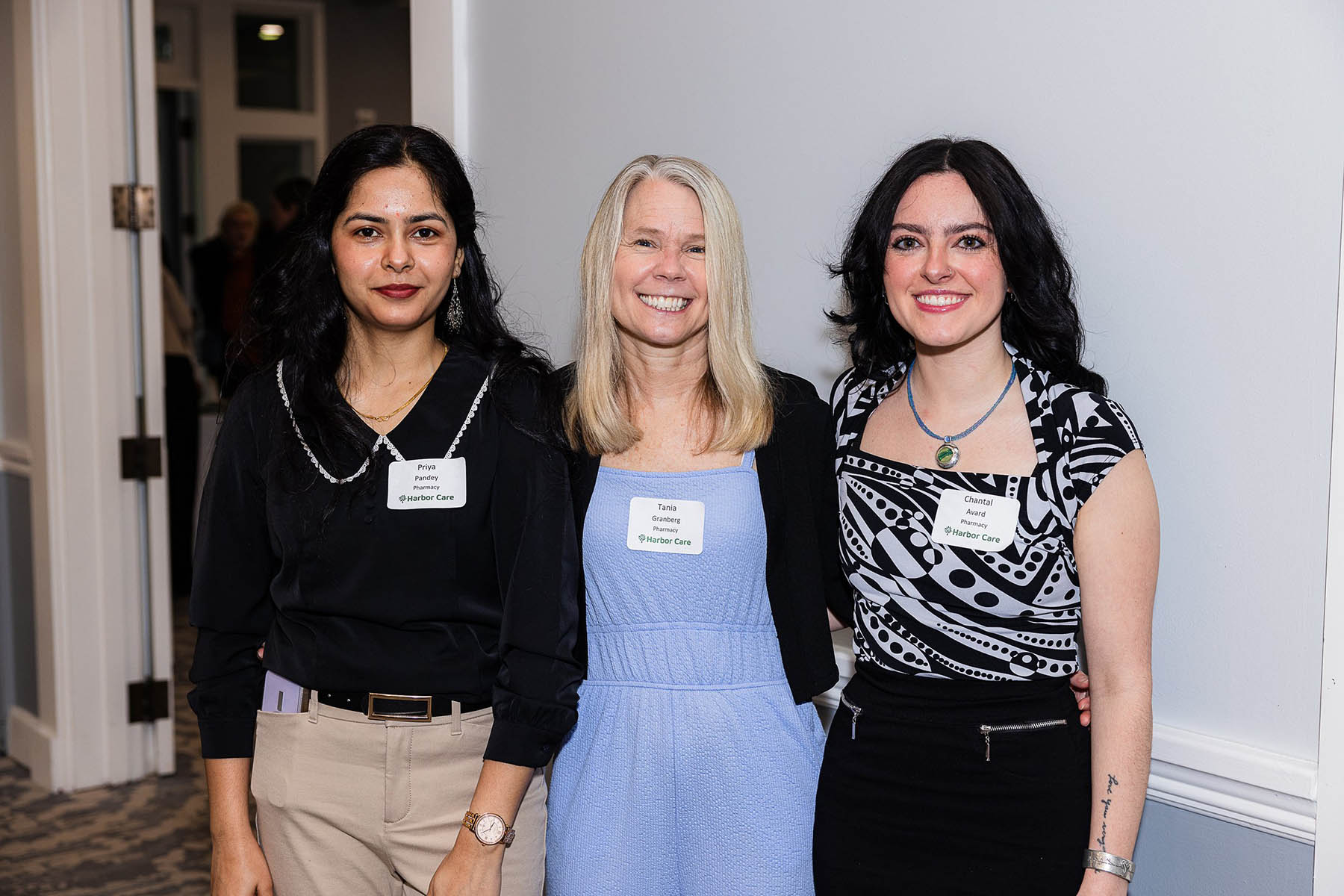 Three people smiling stand side by side at an indoor event, wearing name tags. The person on the left is in a black top, the middle in a light blue dress, and the right in a patterned top and black skirt. The background shows a gray wall and a doorway.