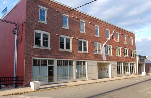 Red brick three-story building with multiple windows and an American flag on a pole. The structure has white accents and a sidewalk in front. The sky is partly cloudy, and the street is empty.