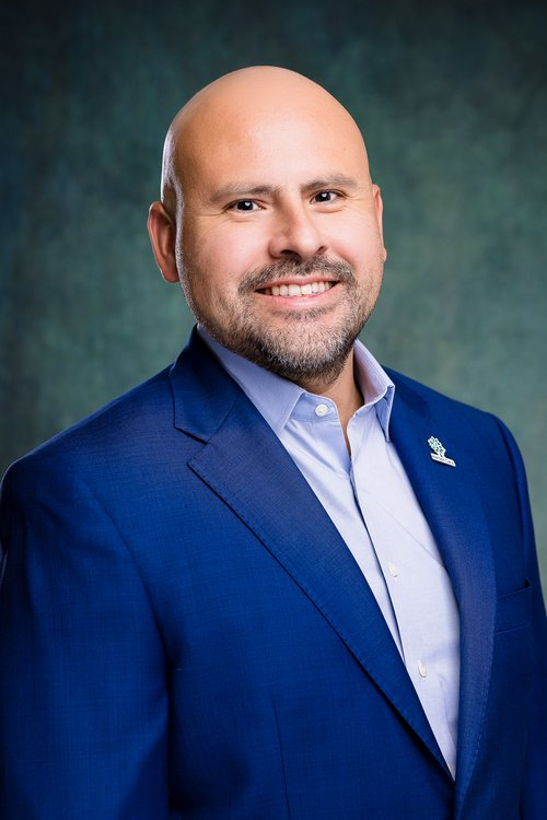 A smiling man in a blue suit and light blue shirt poses against a dark green background. He has a bald head, beard, and is wearing a small lapel pin.