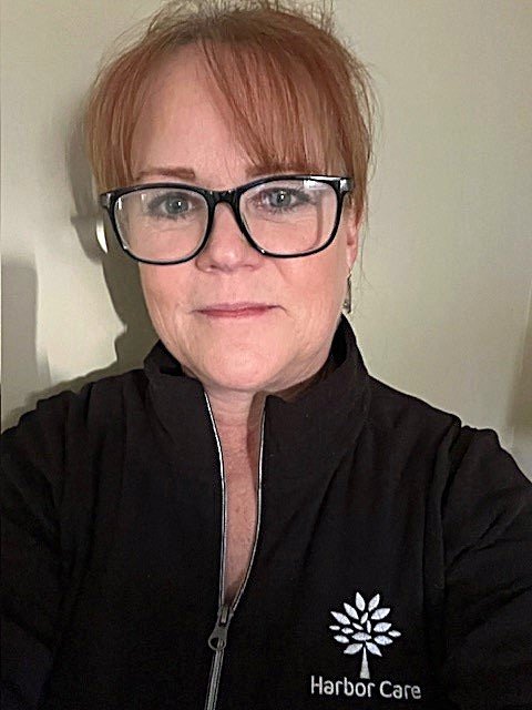 A person with red hair, wearing glasses and a black Harbor Care jacket, is looking at the camera. The logo on the jacket features a white design resembling a flower.