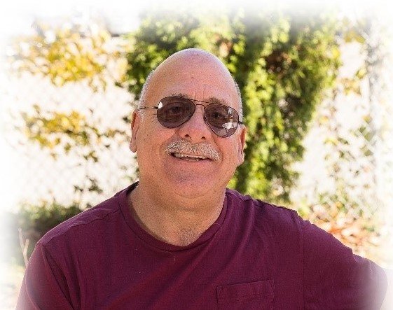 A man with a bald head, mustache, and sunglasses is smiling while wearing a maroon shirt outdoors with greenery in the background.