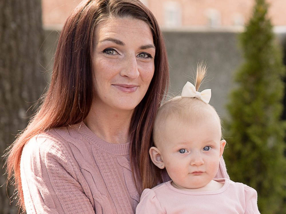 A woman with long brown hair and a baby wearing a pink top sit outdoors. The baby has a small bow in her hair. The background is blurred with trees and a building visible.