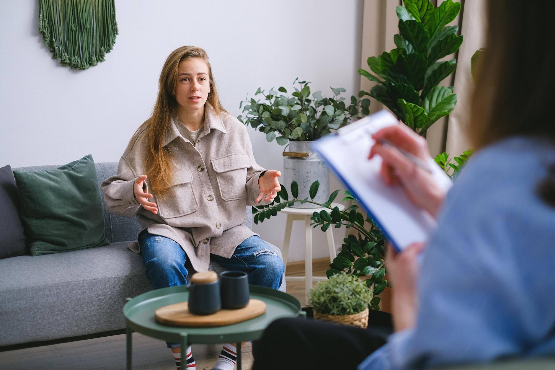 A person with long hair sits on a gray couch, gesturing with hands while talking. They wear a beige jacket and jeans. Another person holds a clipboard in the foreground. The room has plants and a cozy atmosphere.