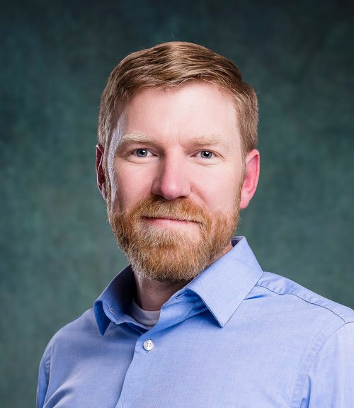A man with a beard and short hair poses against a textured green background, wearing a light blue collared shirt. He looks directly at the camera with a neutral expression.