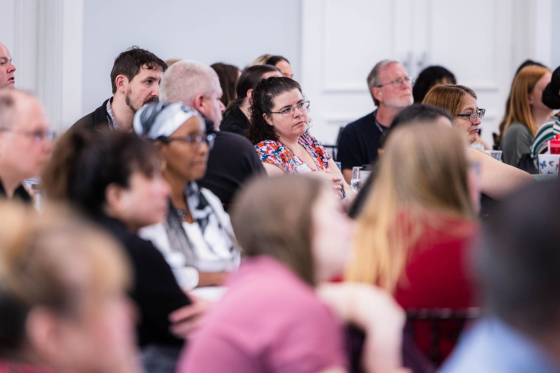 A diverse group of people seated and attentively listening during an indoor event or conference. The focus is on several individuals wearing casual clothing. The background shows a white wall and door.
