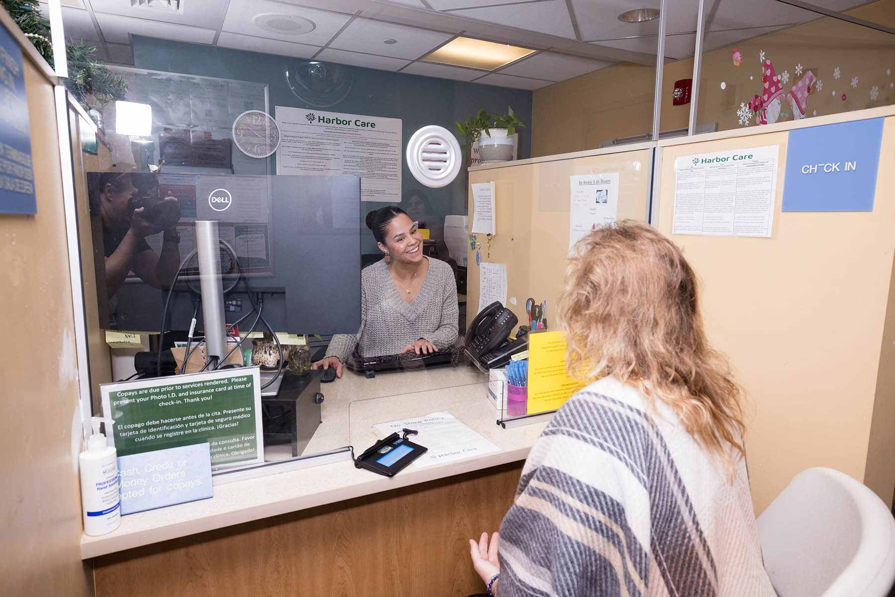 A woman is sitting at a reception desk behind a glass divider, smiling at another woman standing on the opposite side. A photographer is visibly taking pictures from the left. Signs and documents are visible on the walls.