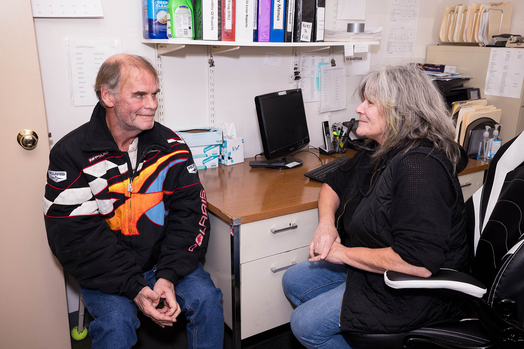 A man and a woman are sitting in an office. The man is wearing a black jacket with colorful patterns, and the woman is in a black vest. They are talking, surrounded by office supplies, a computer monitor, and paperwork on shelves.