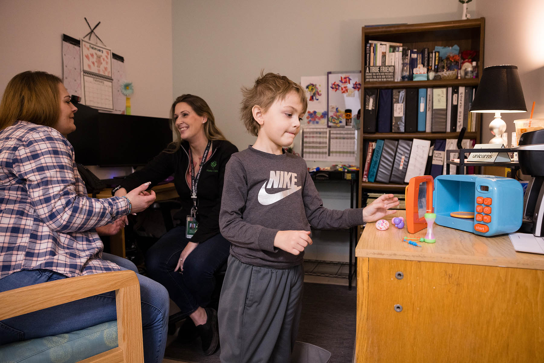 A child plays with a toy cash register on a desk while two adults sit and talk in an office setting.