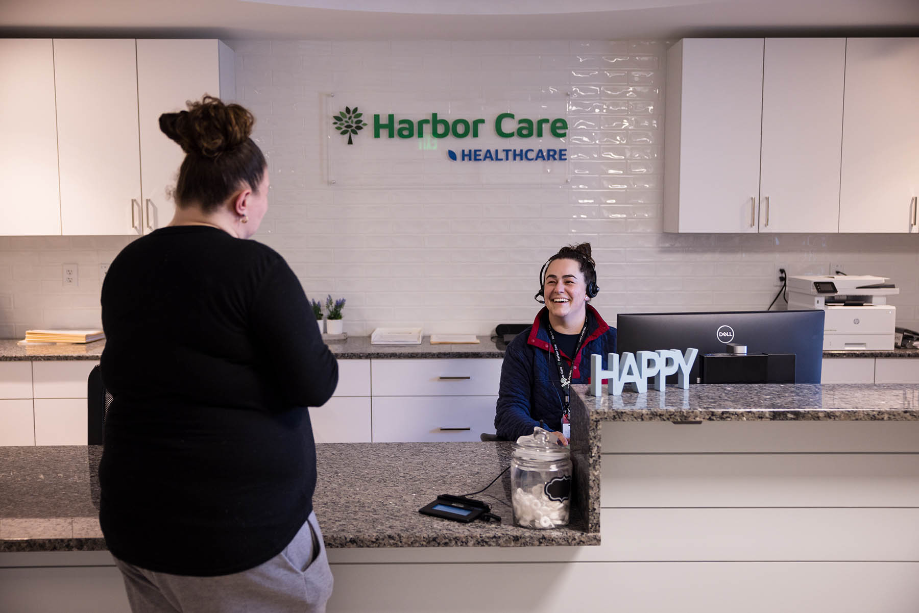 A receptionist wearing a headset smiles while talking to a person standing at the front desk. The desk has a sign that reads HAPPY. The wall behind features the Harbor Care Healthcare logo.