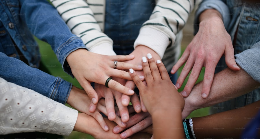 A diverse group of people standing in a circle with their hands layered on top of each other, symbolizing unity and teamwork. Hands show various skin tones, jewelry, and sleeves.