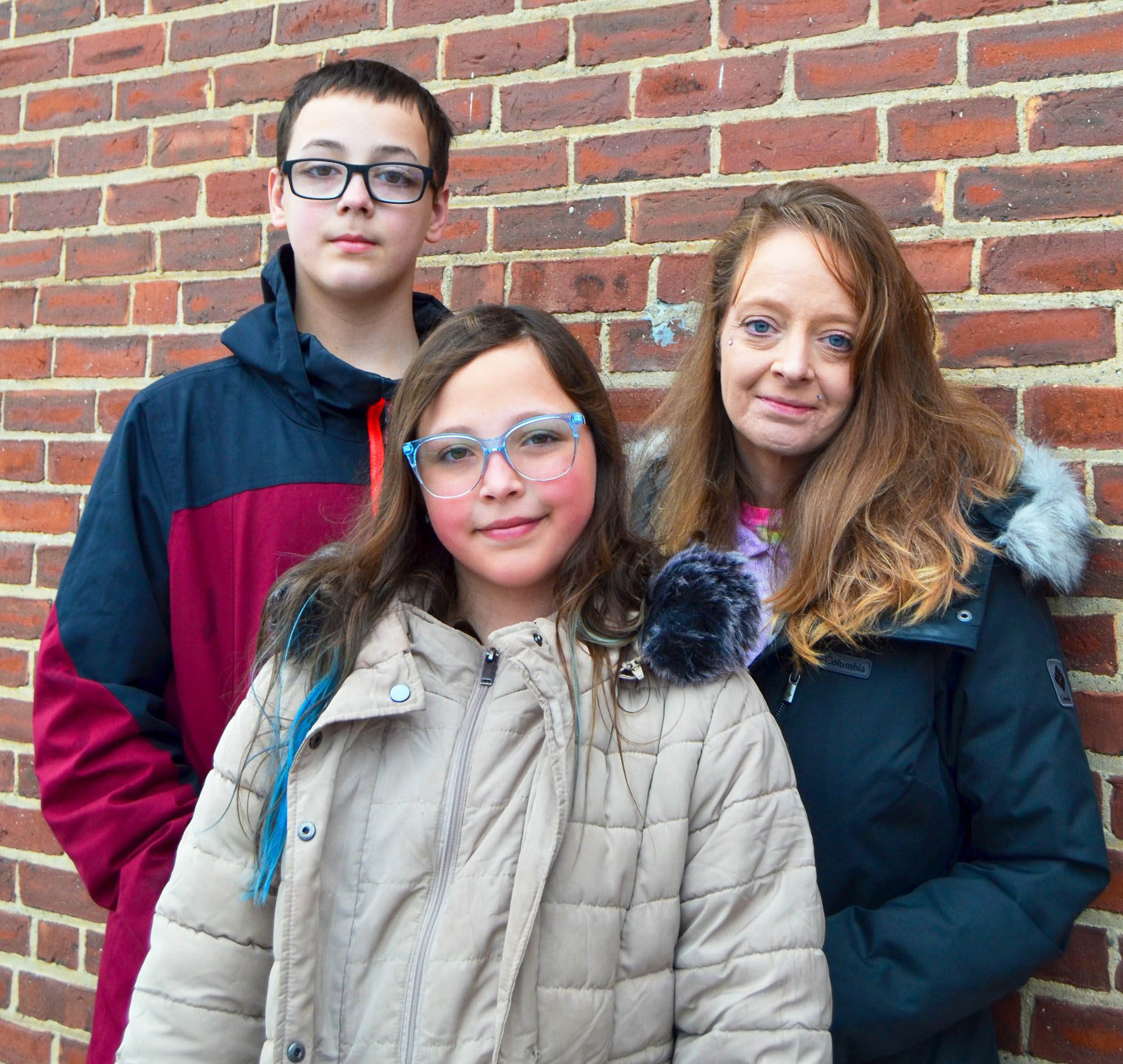 A woman stands with a young girl and boy in front of a brick wall. The girl wears glasses and a beige coat, the boy has short dark hair and glasses with a red and black jacket, and the woman has long hair and a dark coat.