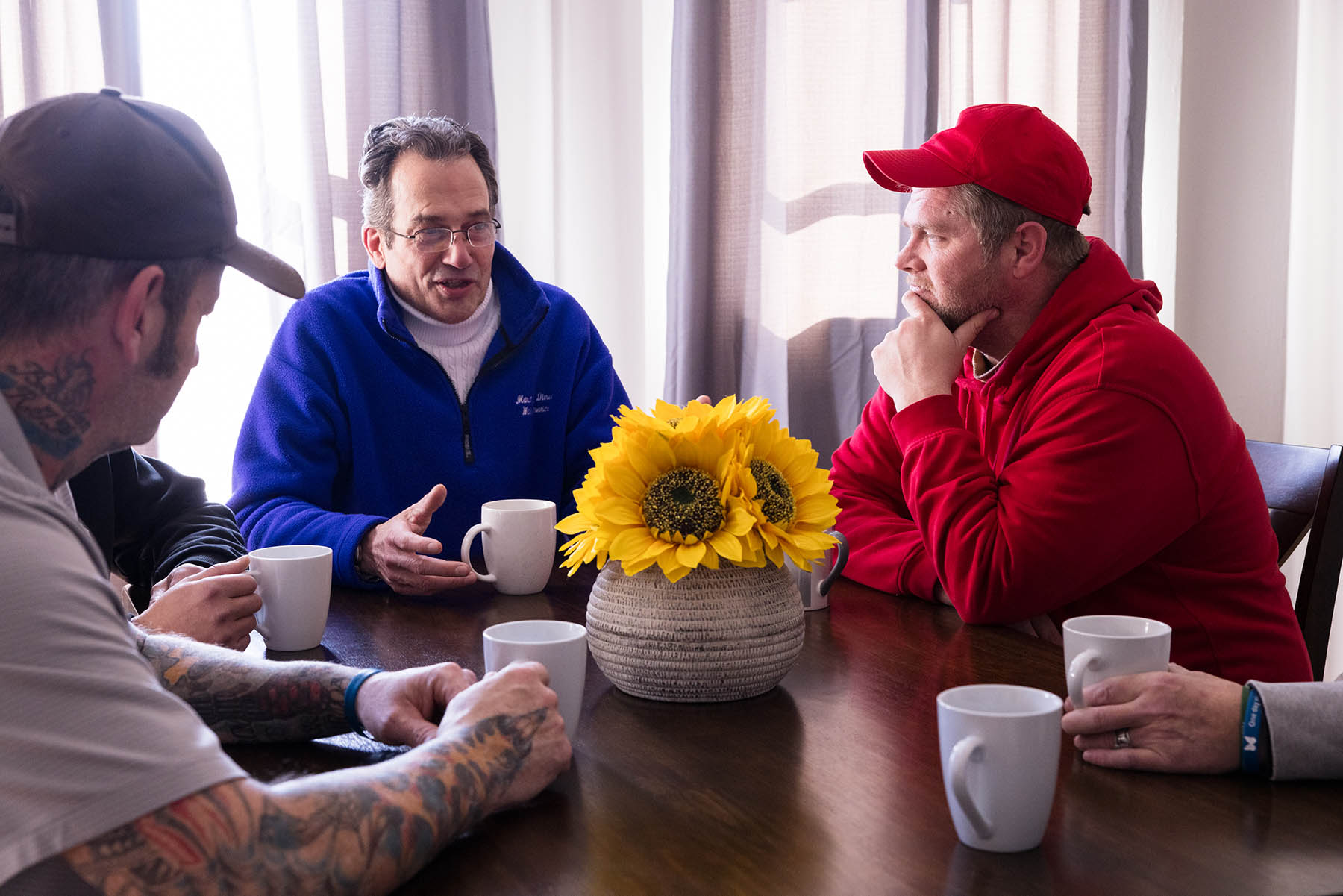 Four men sit around a table with coffee mugs, engaged in conversation. A vase with sunflowers is placed at the center of the table.