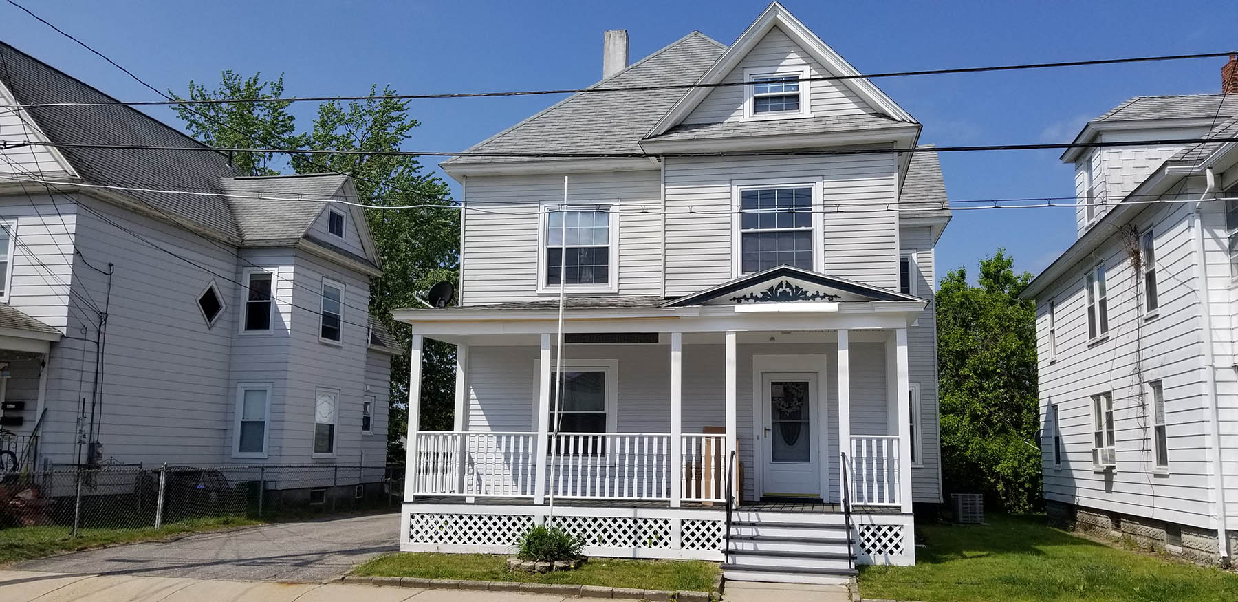 A two-story white house with a covered front porch, black trim, and a gabled roof, situated between similar neighboring homes on a sunny day.
