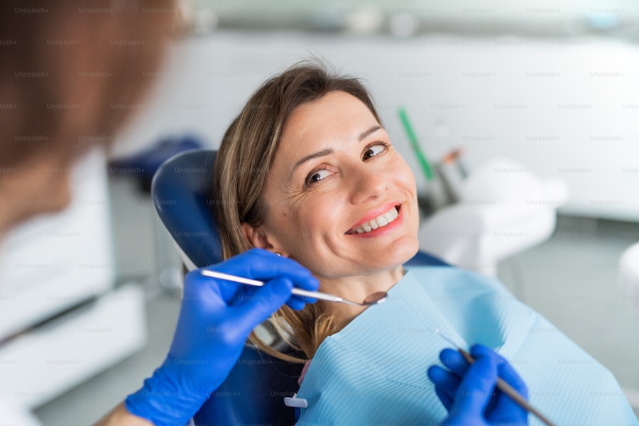 A woman in a dental chair smiles at the camera while a dentist wearing blue gloves examines her teeth with dental tools. She is wearing a blue dental bib. The background is slightly blurred, showing dental equipment.