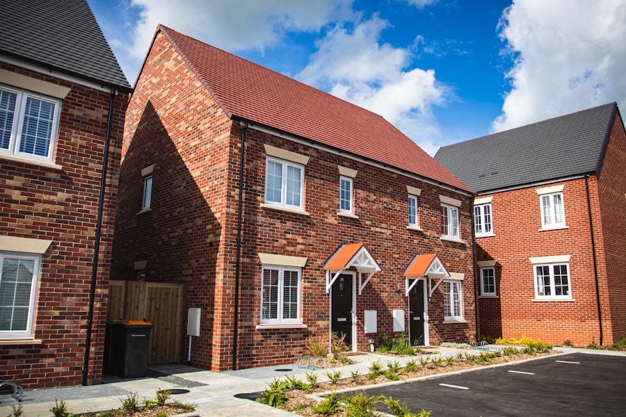 A row of modern brick houses with pitched roofs and white-framed windows under a partly cloudy sky. The front area features small garden plots and a paved walkway leading to black front doors.