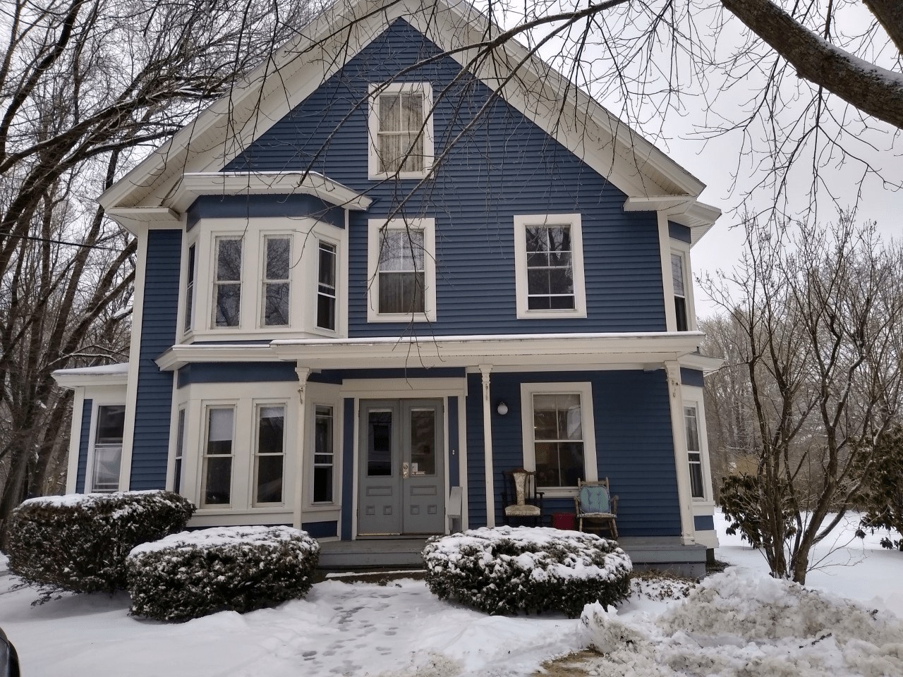 A blue two-story Victorian-style house with white trim stands in a snowy landscape. There are several windows, a covered porch, and snow-covered shrubs in the front. Leafless trees surround the house, and the sky is overcast.