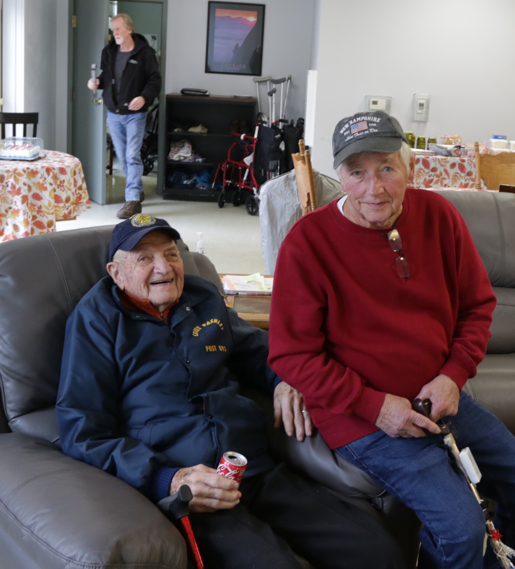 Two elderly men, both wearing caps and seated on a couch, pose for the camera while another man stands in the background near tables with tablecloths.
