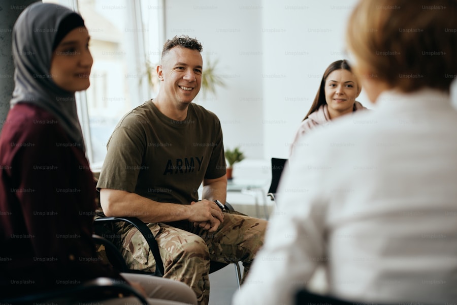 A diverse group of people sitting in a circle indoors, engaged in conversation. A man in military attire smiles, while others listen attentively. The setting suggests a supportive or group discussion environment.