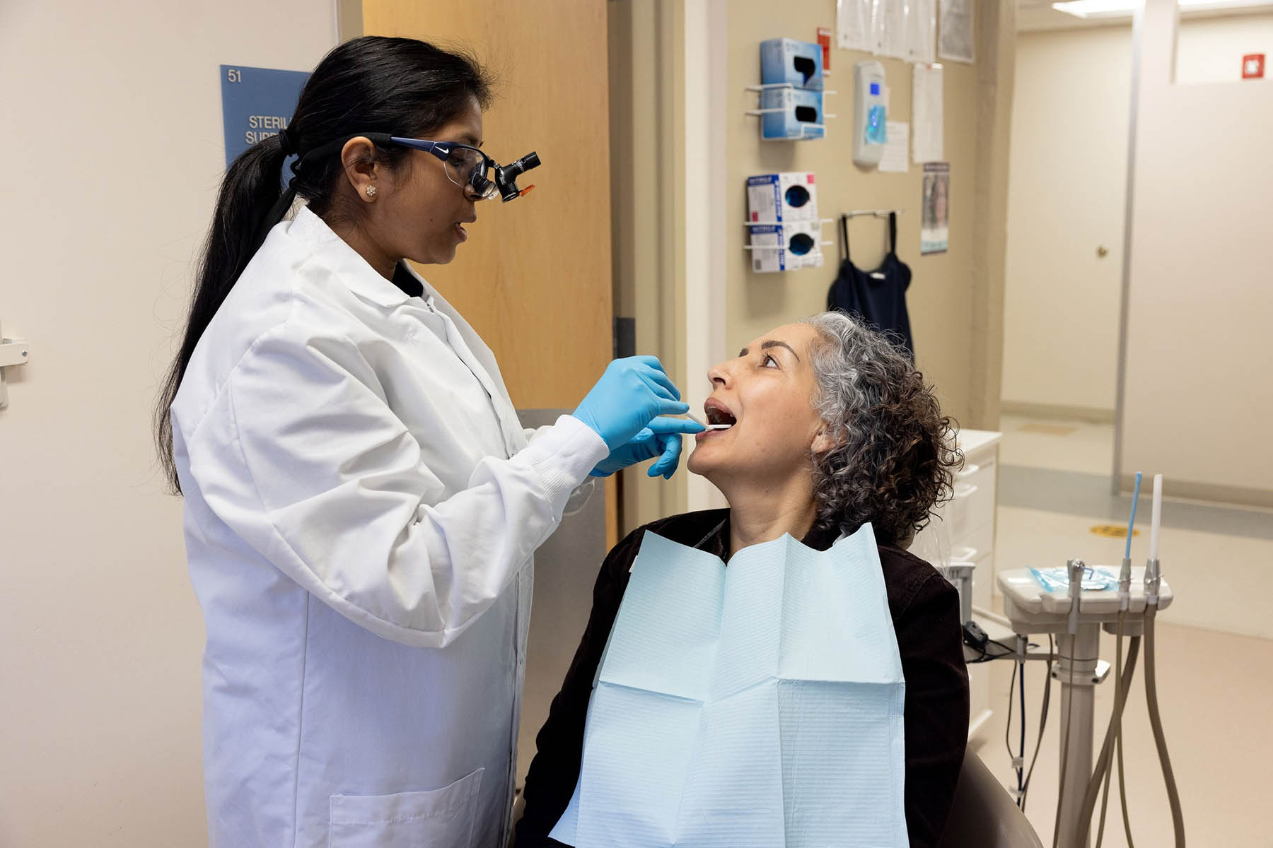 A dentist wearing magnifying glasses and gloves examines a patients teeth in a dental clinic. The patient, seated and wearing a bib, looks up at the dentist. Dental equipment and supplies are visible in the background.