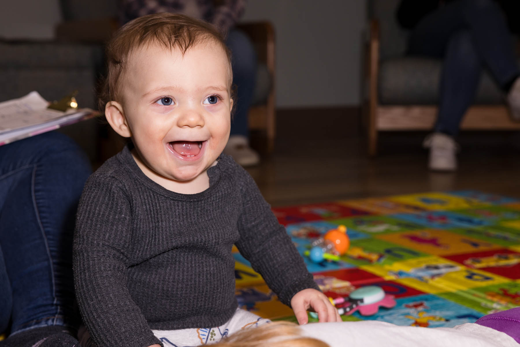 non-printable A baby in a dark sweater is sitting on a colorful play mat, smiling widely. Toys are scattered in the background, and adults are seated behind.