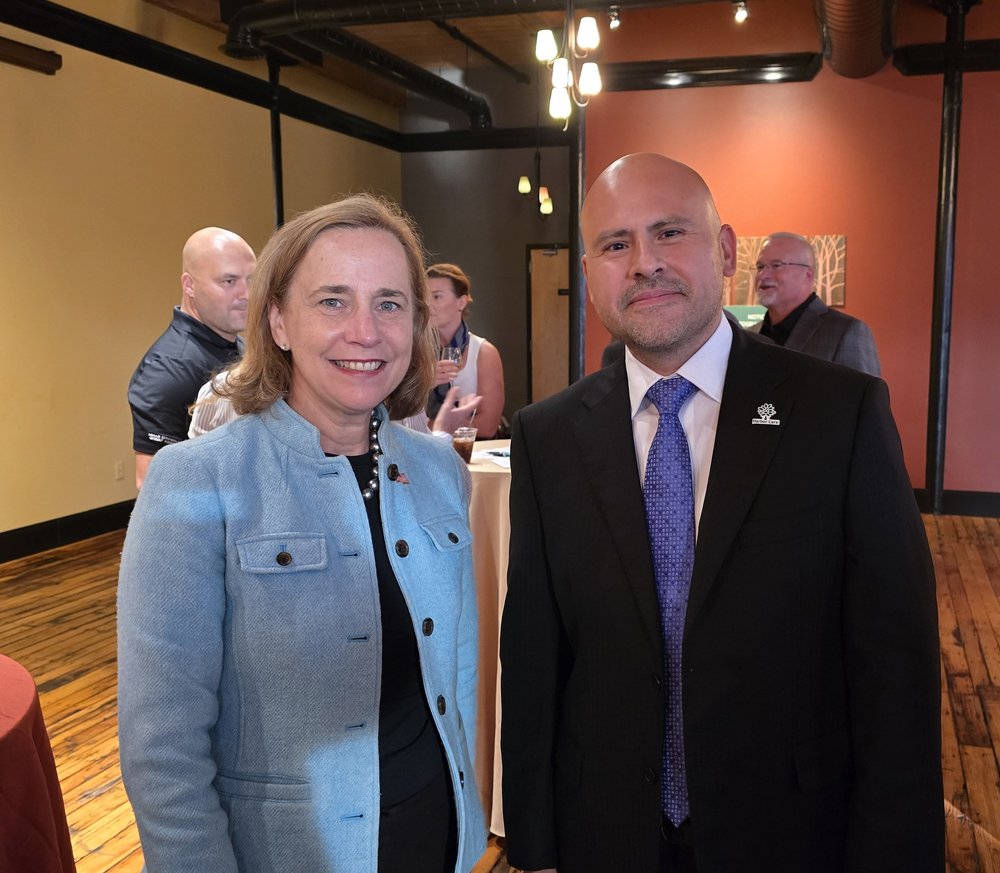 Two individuals are standing side by side in a warmly lit room with wooden floors and earth-toned walls. The person on the left wears a light blue jacket, while the person on the right is in a suit and tie. Others are visible in the background.