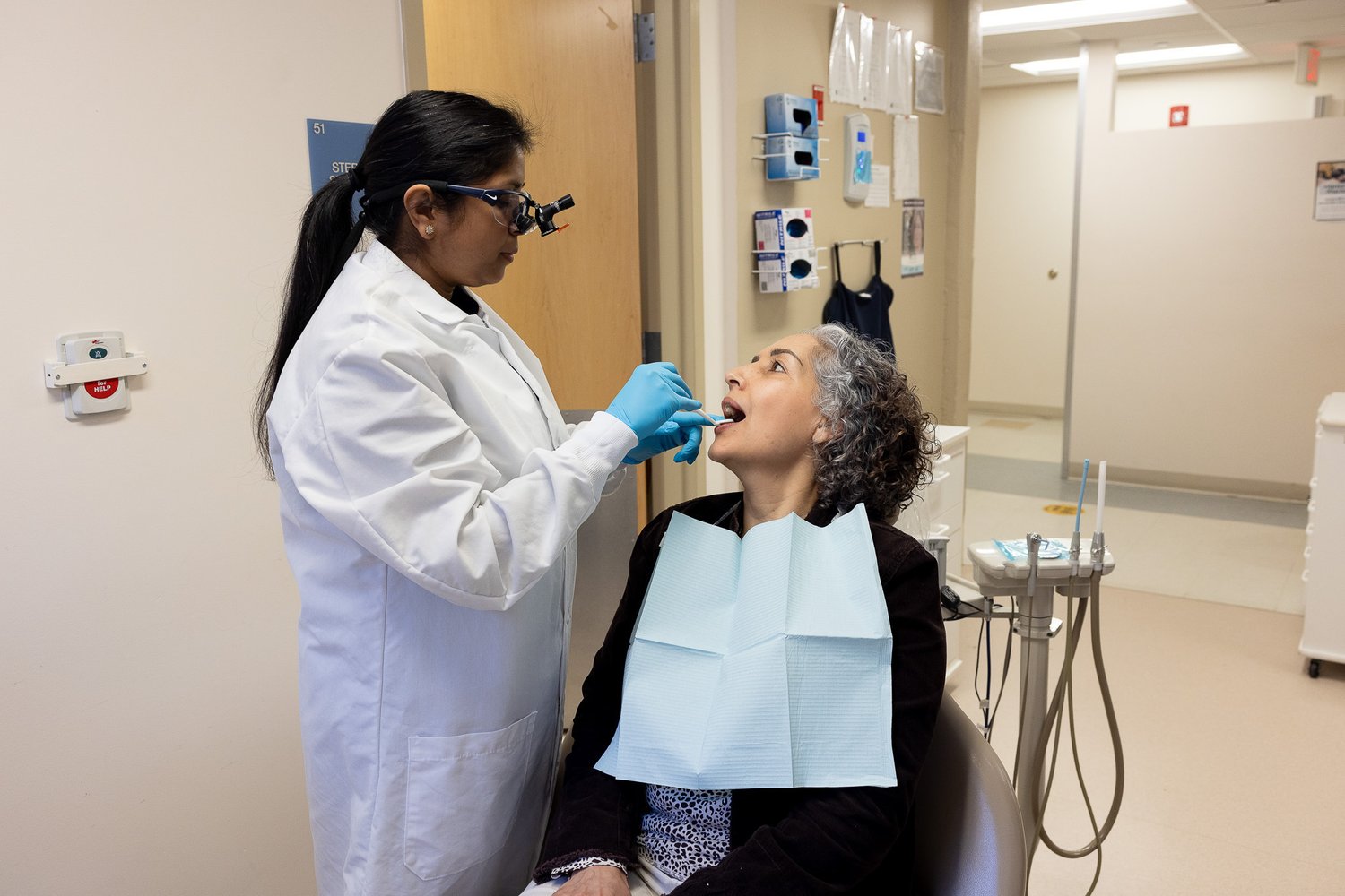 A dentist wearing a white coat and magnifying eyewear examines a patients mouth. The patient sits in a dental chair with a bib. Dental tools are visible nearby in the office.