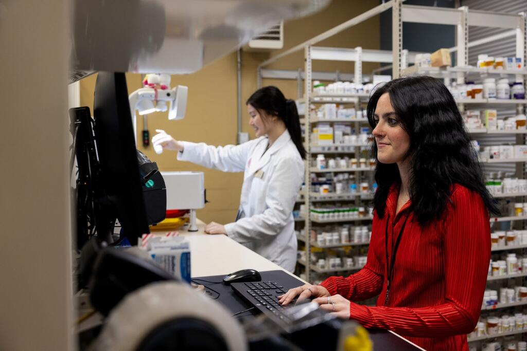 Two pharmacists working in a pharmacy. The woman in the foreground, wearing a red blouse, is using a computer. Behind her, another pharmacist in a white coat is organizing medicine on shelves. The shelves are filled with various medication bottles.