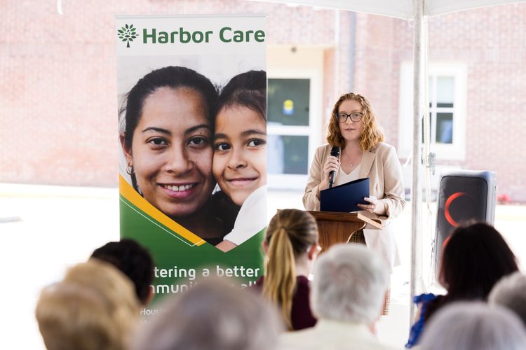 A woman stands at a podium speaking into a microphone at an outdoor event. Beside her, a poster displays two smiling faces and the text Harbor Care: Fostering a better community.