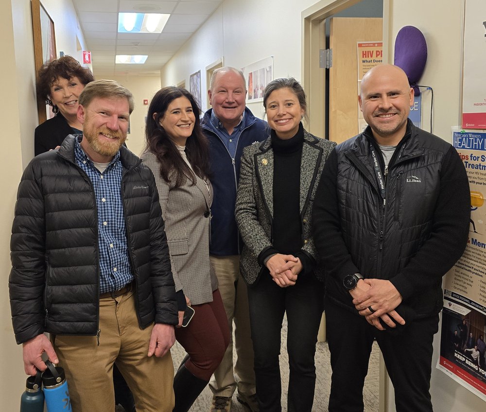 Six adults stand and smile together in a hallway lined with posters and information on the walls.