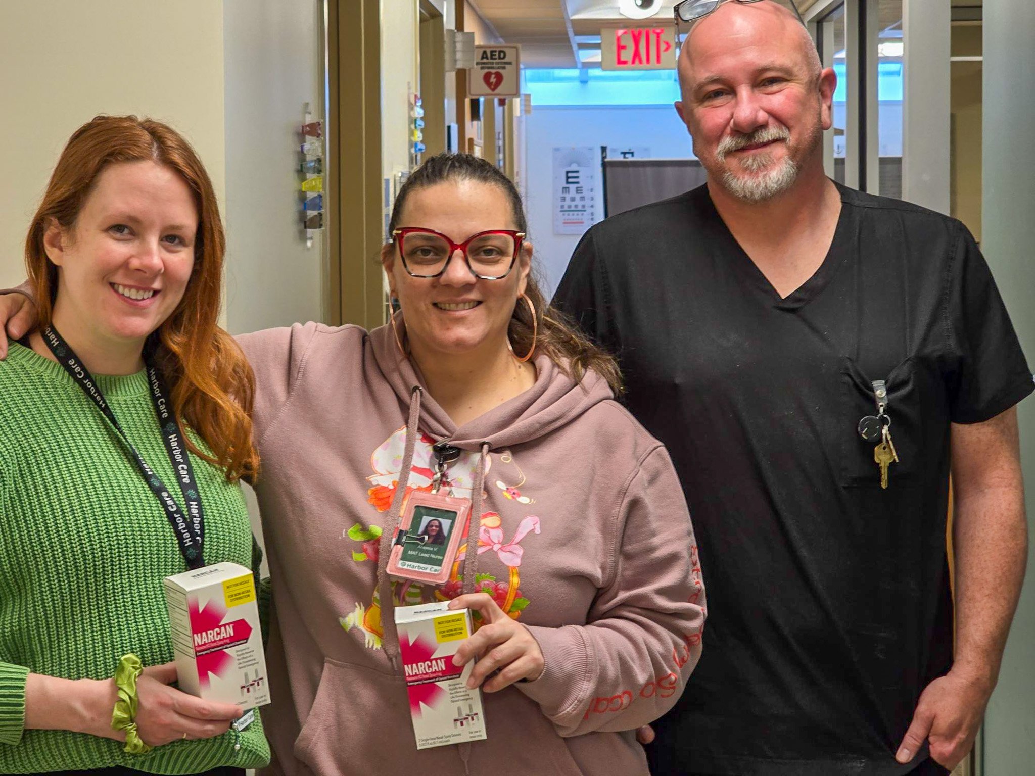 Three people stand indoors, smiling at the camera. Two of them hold boxes of Narcan nasal spray.