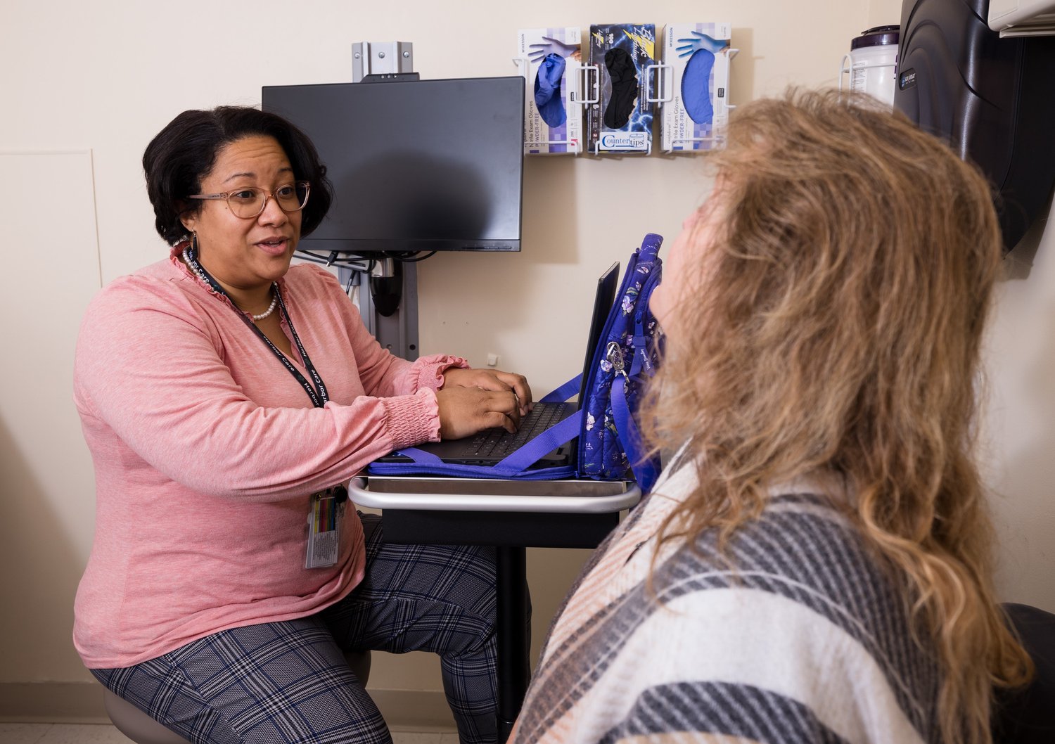 A healthcare professional in a pink sweater sits at a desk with a laptop, speaking to a patient wrapped in a plaid blanket. Medical supplies are visible on the wall in the background.