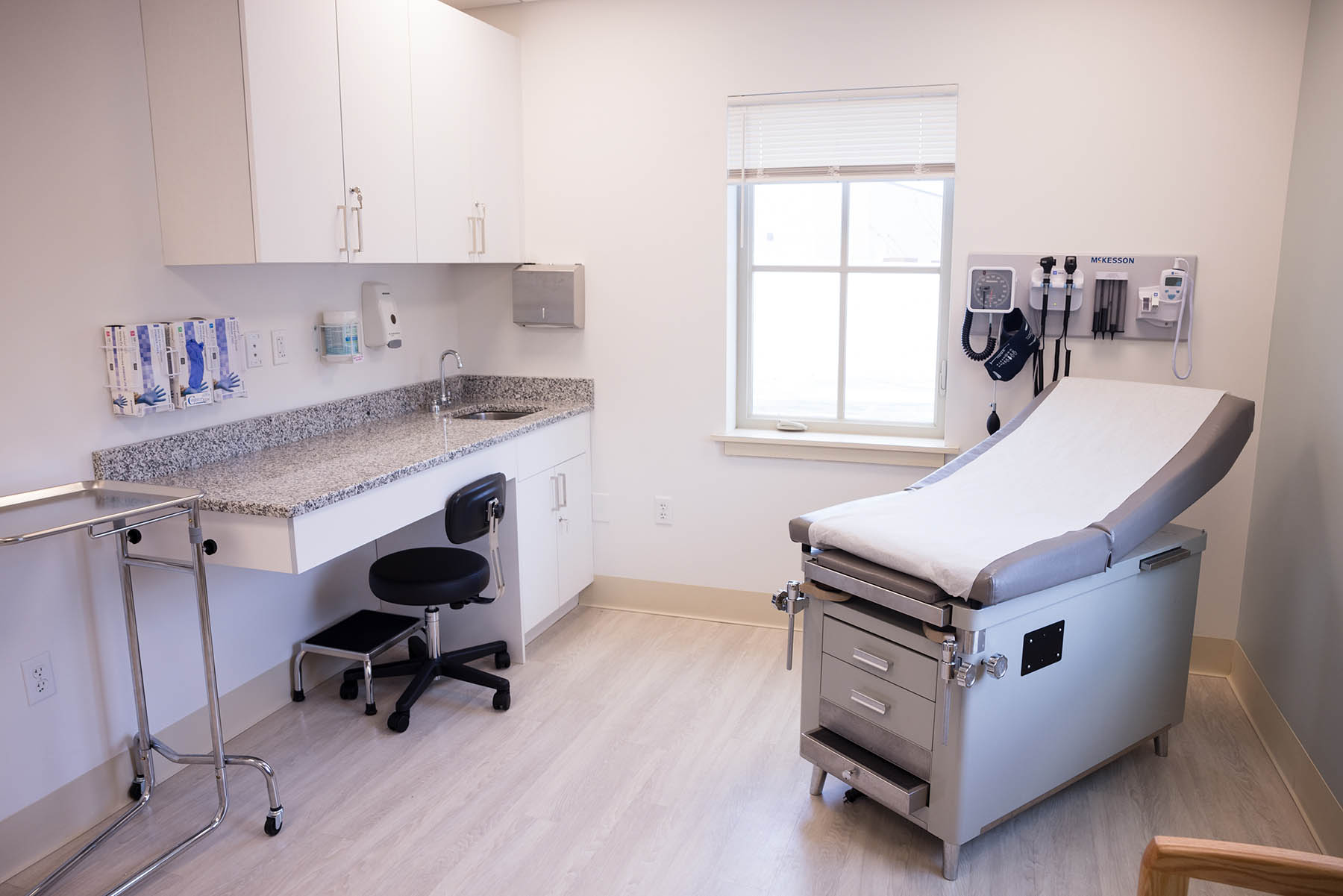 A clean, bright medical examination room with an exam table, a sink with cabinets above, a stool, a tray table, and medical equipment on the wall. A window allows natural light to enter the room.