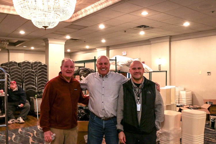 Three men standing together in a room with a chandelier. They are smiling and posing for the photo. Behind them, there are stacks of chairs, shelves with bags, and various items scattered around. The room has a carpeted floor and ceiling lights.