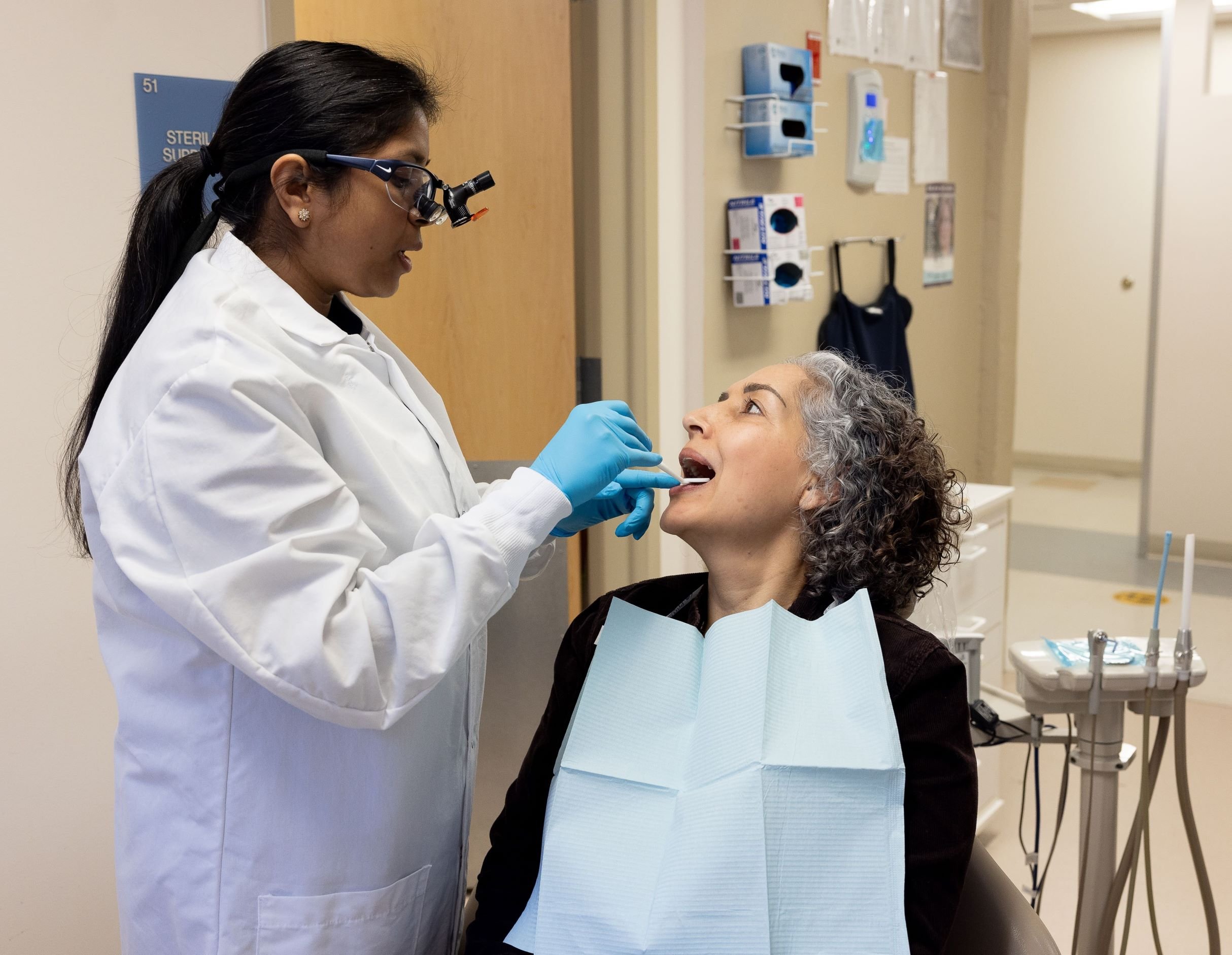 A dentist in a white coat and magnifying glasses examines a patients mouth. The patient, wearing a dental bib, is seated and has curly gray hair. Dental tools and supplies are visible in the background.