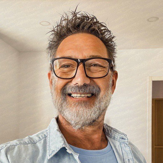 A middle-aged man with glasses, gray hair, and a beard smiles at the camera in a brightly lit room.