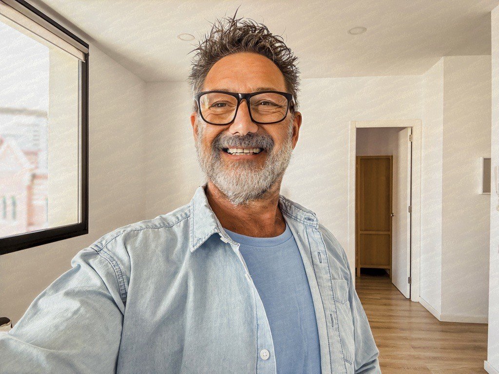 Man with glasses and a beard smiles while taking a selfie indoors. He is wearing a light denim shirt over a blue t-shirt. The room has wooden flooring, a closed door, and a large window letting in natural light.