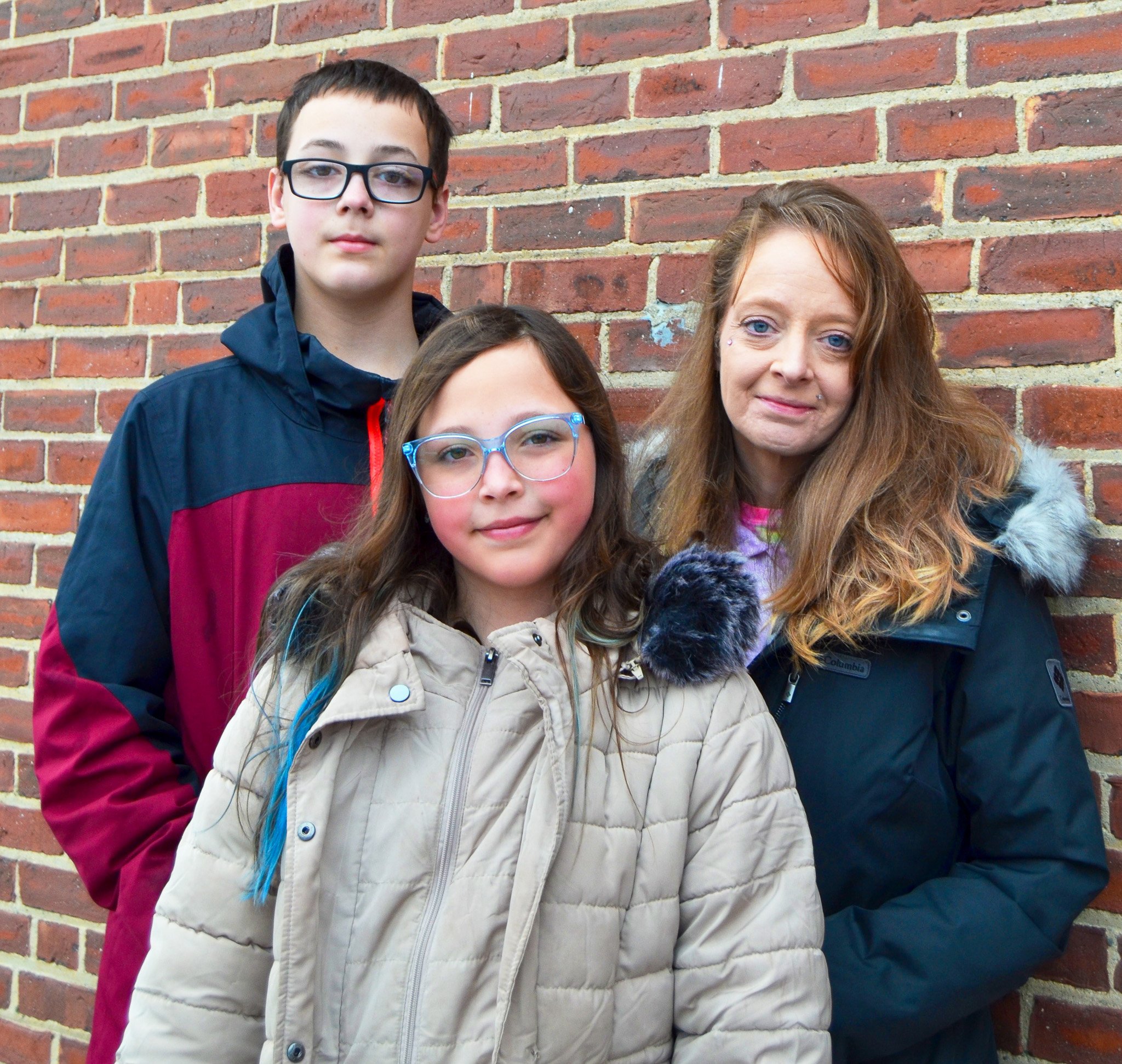A woman and two kids stand in front of a brick wall. The girl in front is wearing a light-colored coat and glasses. The boy is wearing glasses and a jacket. The woman is in a dark coat, with long hair. All are looking at the camera.