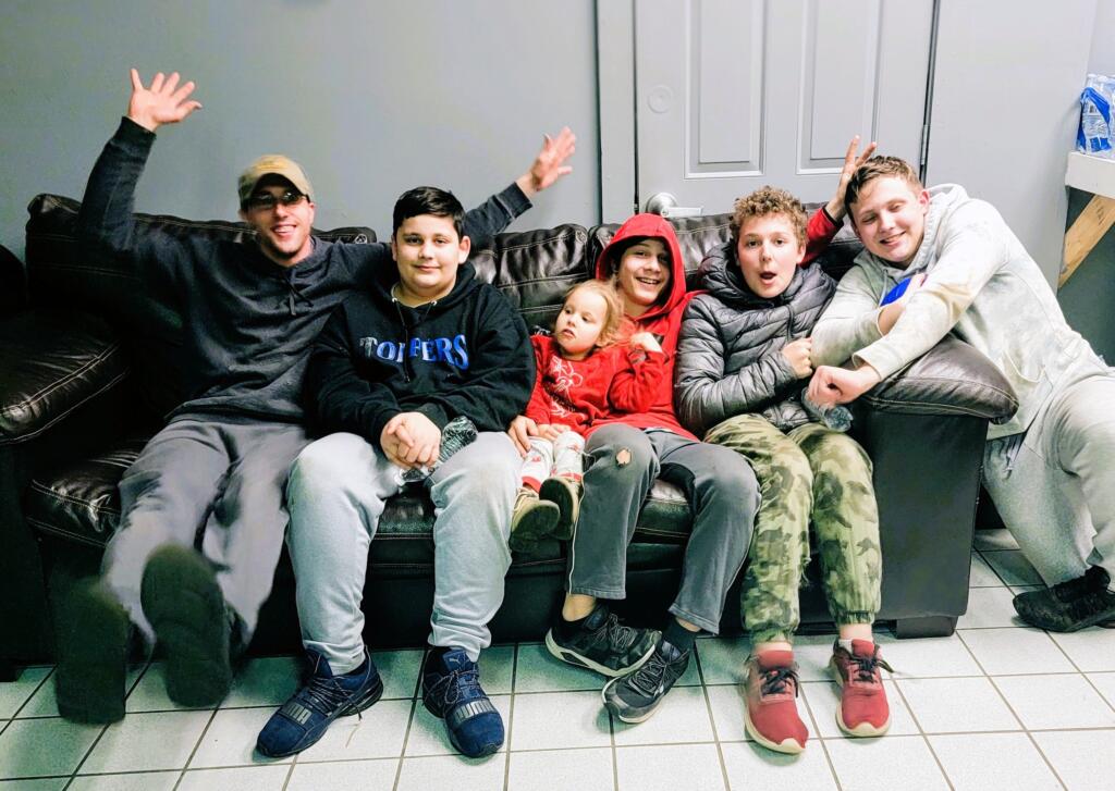 A group of six people, including five boys and a girl, sit on a couch. They are dressed casually, with some wearing hoodies and jackets. One boy has his arms raised playfully, and the girl, in a red hoodie, has her eyes closed. They appear happy and relaxed.