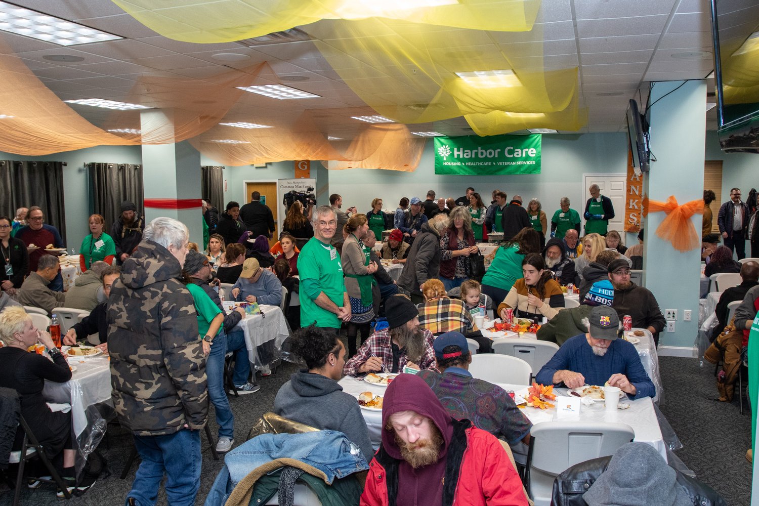 A large group of people sit and stand at tables in a decorated indoor space, eating and socializing during a community meal event. Volunteers in green shirts assist guests; autumn decorations hang overhead.