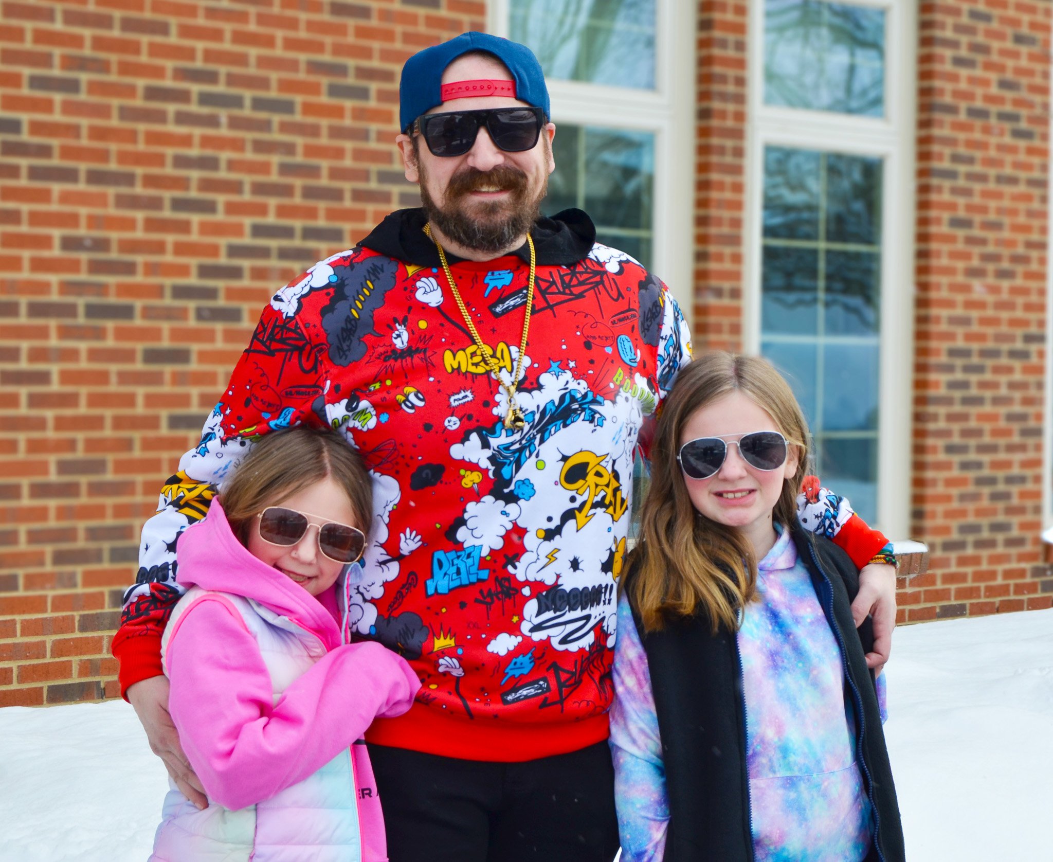 A man wearing a colorful, graphic sweatshirt and sunglasses stands smiling with two young girls in sunglasses, both in winter clothing. They are outside, with a brick building and snow in the background.