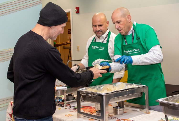 Two men in green aprons serve food to a person in a black sweater and hat. They are standing behind a table with serving trays filled with food. The scene appears to be in a community or cafeteria setting.