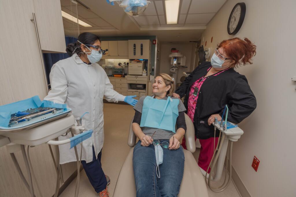 A woman sits in a dental chair smiling at two dental professionals standing nearby, all wearing masks and scrubs, in a modern dental clinic. Dental equipment is visible around them.