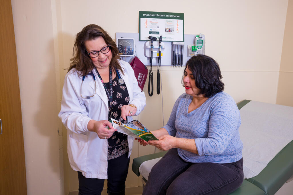 A doctor stands next to a seated patient in a medical exam room, both looking at a brochure together. Medical equipment is mounted on the wall behind them.