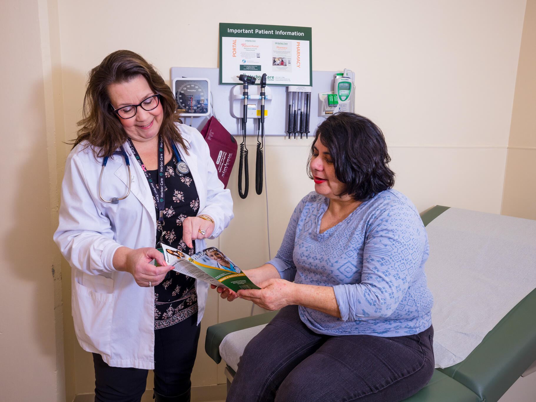 A doctor stands next to a seated patient in a medical exam room, both looking at a brochure together. Medical equipment is mounted on the wall behind them.