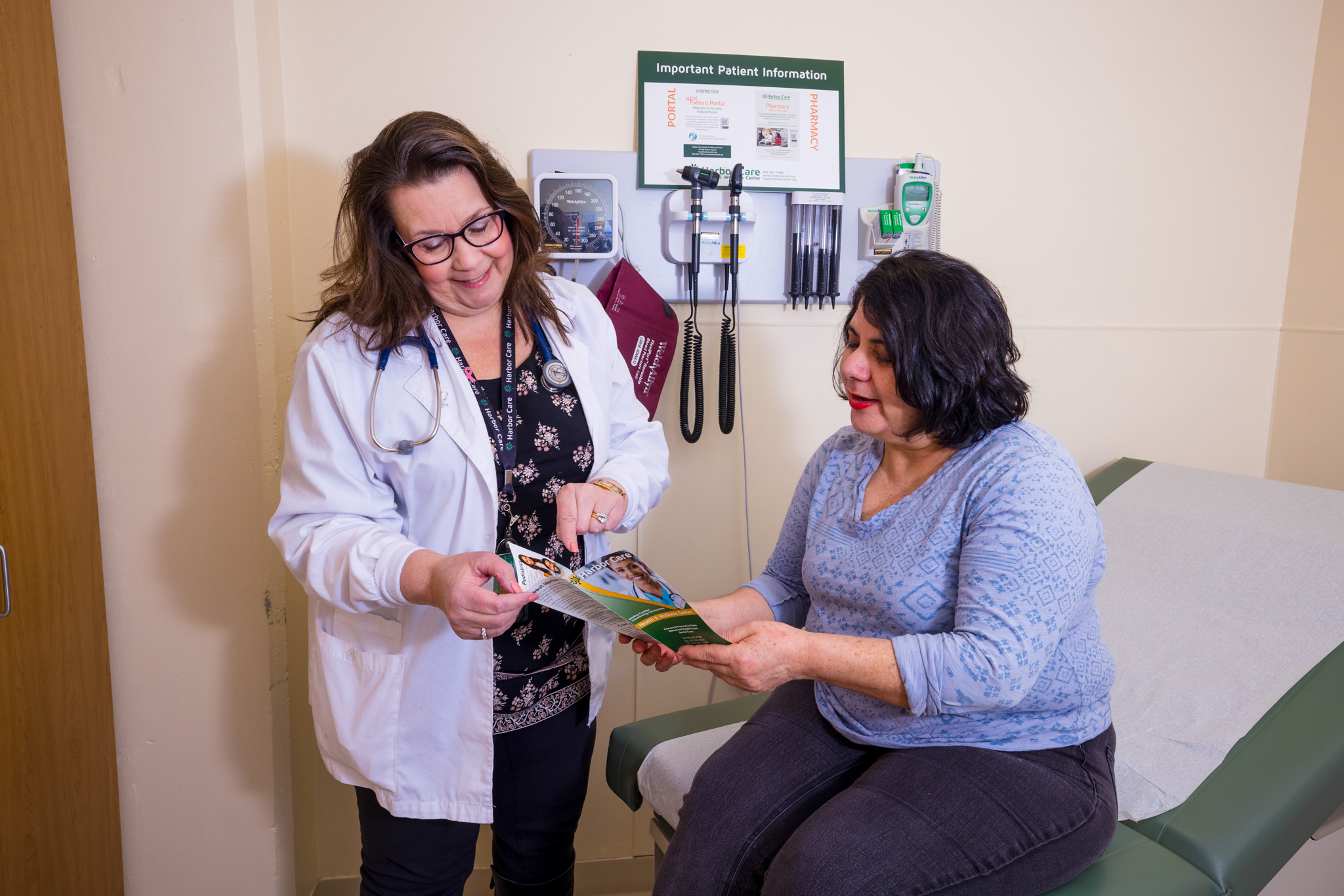 A doctor stands next to a seated patient in a medical exam room, both looking at a brochure together. Medical equipment is mounted on the wall behind them.
