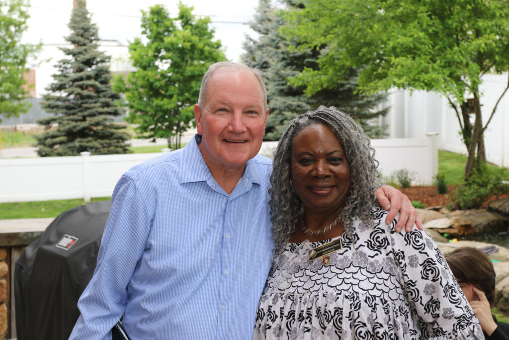 Two older adults, one man and one woman, stand close together outside, smiling at the camera with trees and a white fence in the background.