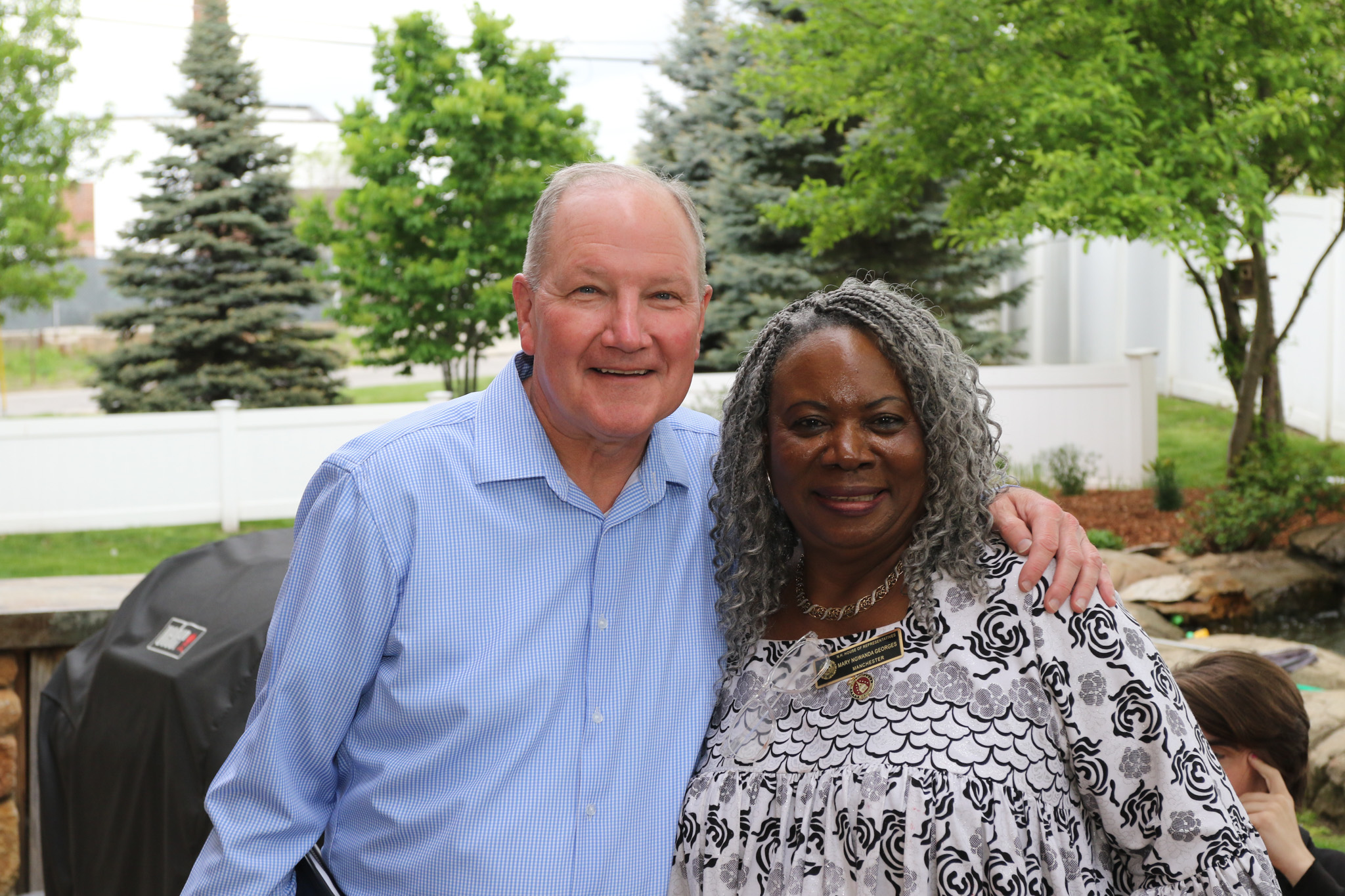 Two older adults, one man and one woman, stand close together outside, smiling at the camera with trees and a white fence in the background.
