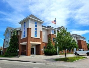 BAE+Systems+Independence+Hall+housing+Manchester A modern three-story brick and white building with large windows, surrounded by trees. An American flag on a pole stands in front, and a white truck is parked nearby under a partly cloudy sky.