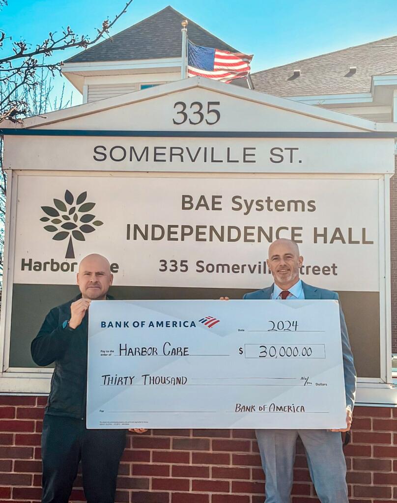 Two men stand in front of a building sign holding a large ceremonial check for $30,000 made out to Harbor Care from Bank of America.